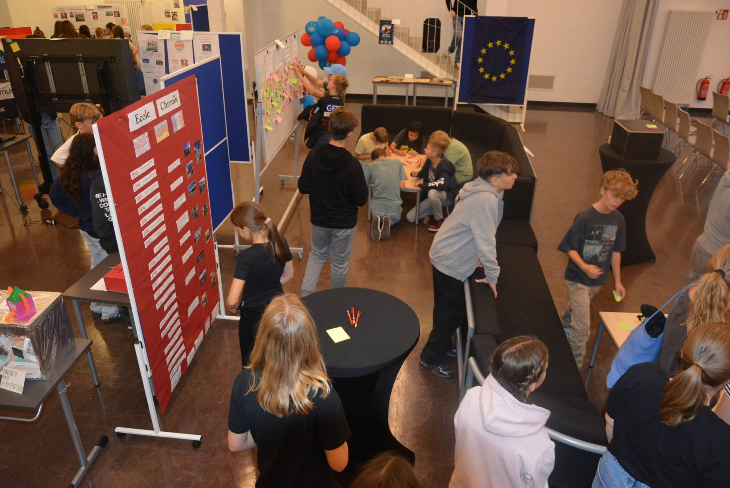 Eine Gruppe von Schülern arbeitet daran, farbige Notizen an Pinnwände zu heften; Poster und EU-Flagge im Hintergrund.