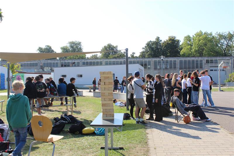 Eine Gruppe von Jugendlichen spielt Jenga im Freien und sitzt auf Bänken. Viel Grün und Sonne.