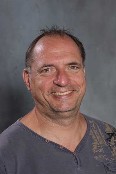 Ein lächelnder Mann mit kurzen Haaren in grauem Hemd vor einem grauen Hintergrund.