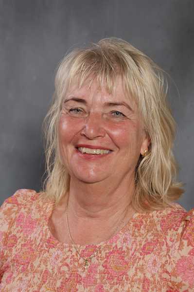 Eine lächelnde Frau mit blonden Haaren und einem rosa gemusterten Oberteil vor grauem Hintergrund.