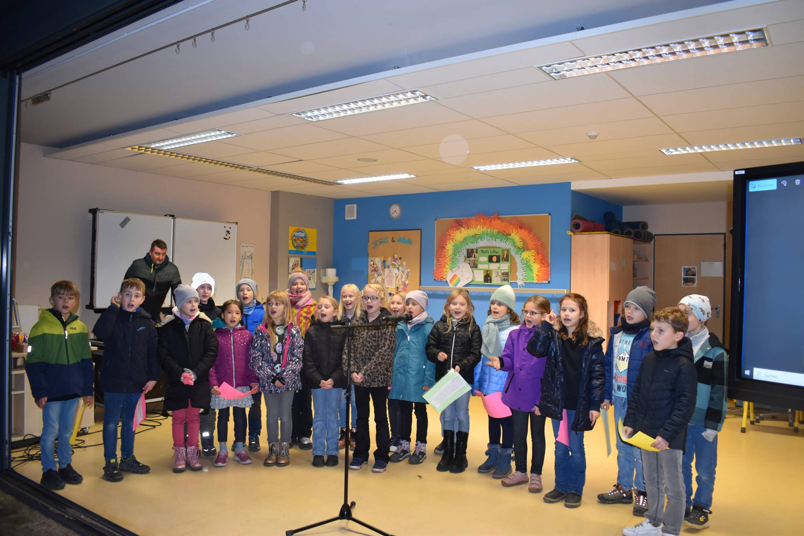 Kinderchor singt freudig; bunte Kleidung, Wandgemälde im Hintergrund, Mikrofon vorn.