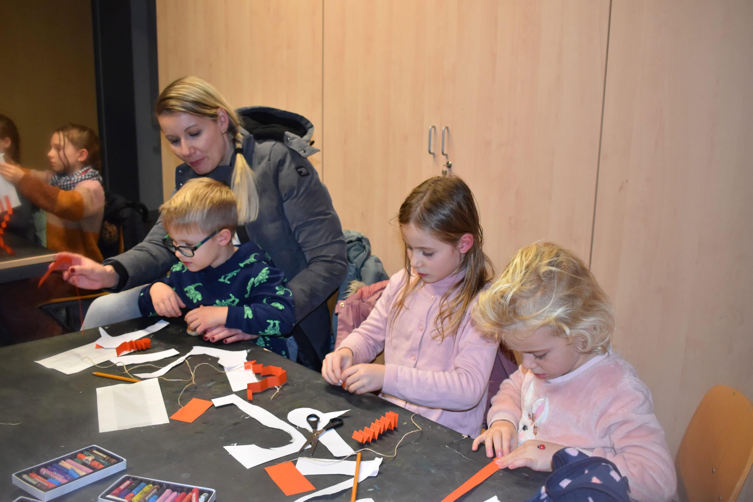 Eine Frau mit drei Kindern bastelt konzentriert mit buntem Papier an einem Tisch.