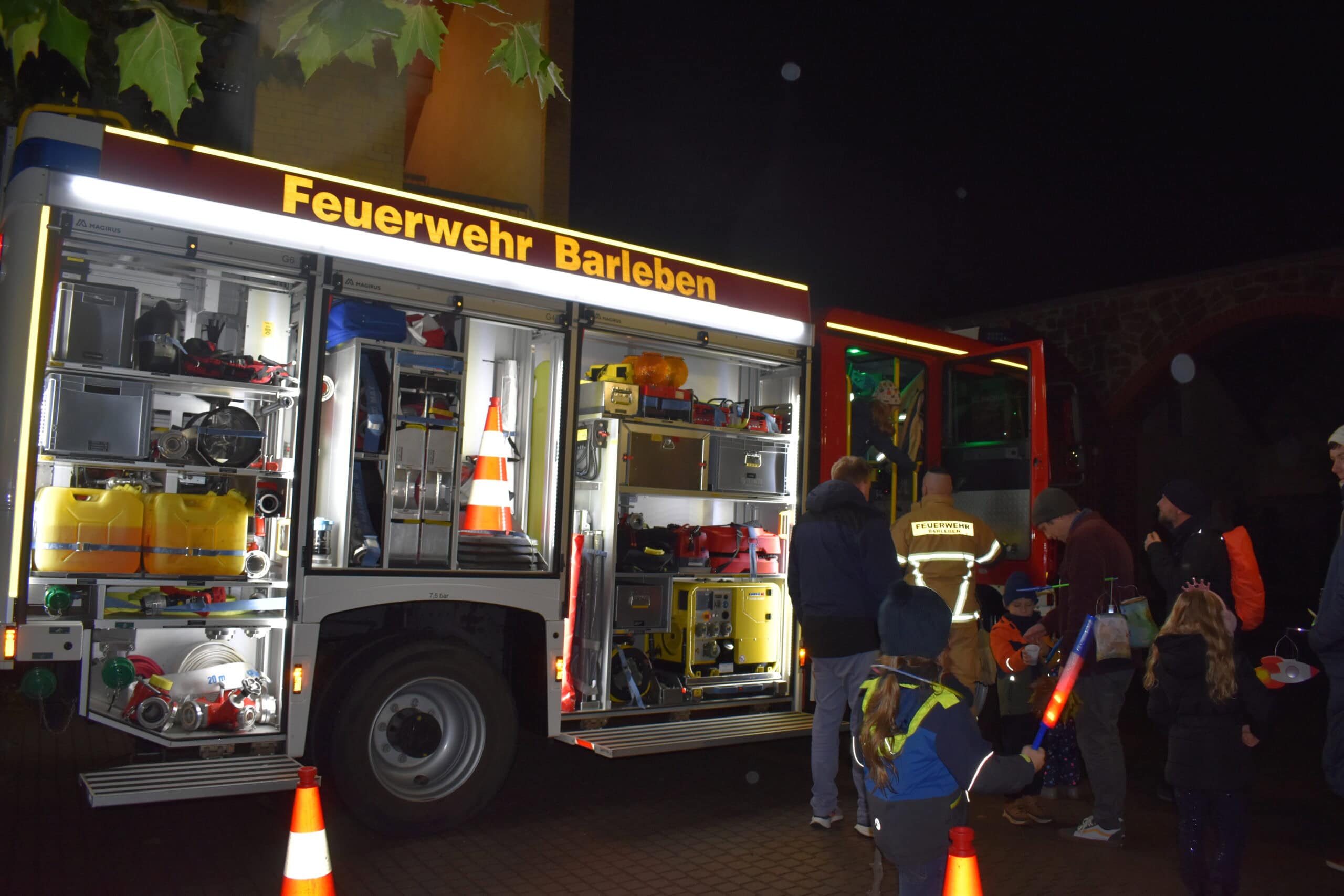 Eine Feuerwehr und Menschen bei Nacht, helle Lichter, reflektierende Westen und orangene Mützen.