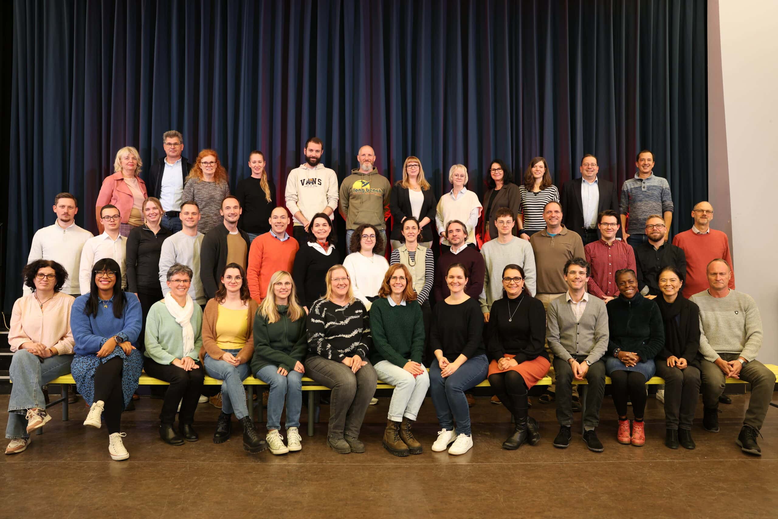 Gruppenfoto von lächelnden Personen vor blauem Vorhang, in bunter Alltagskleidung.