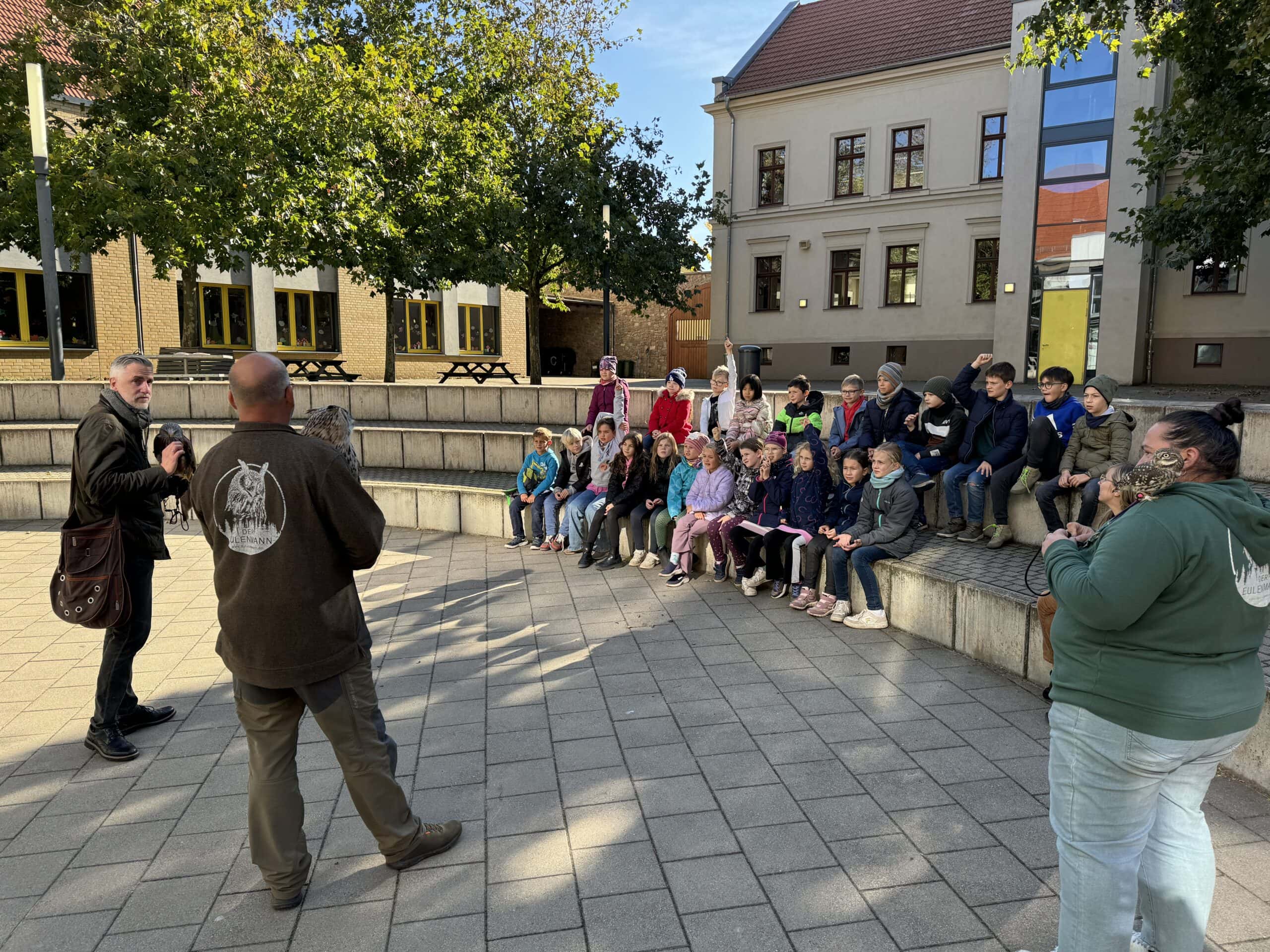 Männer instruieren Kindergruppe im Freien; alle schauen aufmerksam, Eule auf dem Arm eines Mannes.