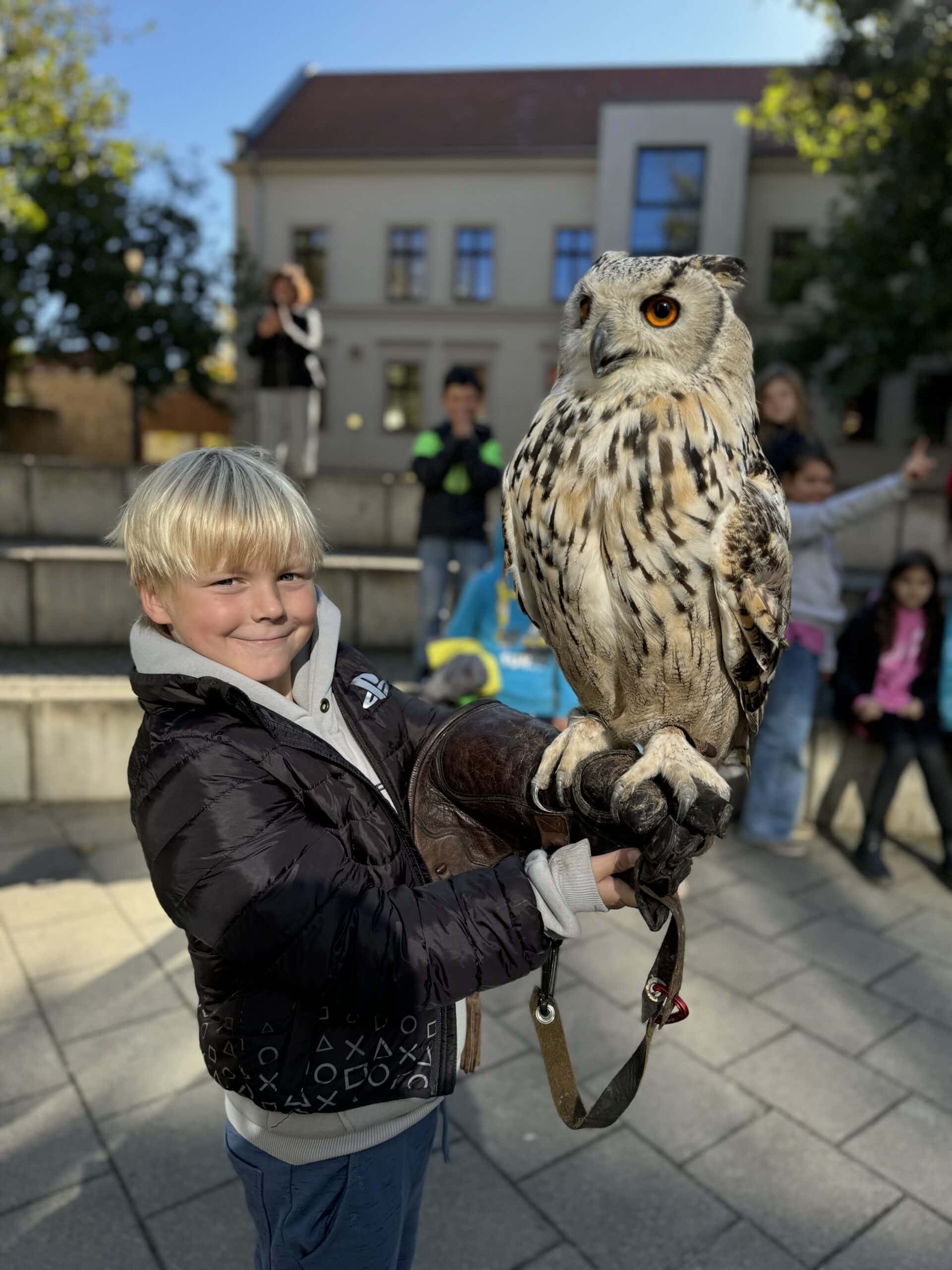 Ein lächelnder Junge hält einen großen Uhu auf einem Platz vor einem Gebäude bei sonnigem Wetter.