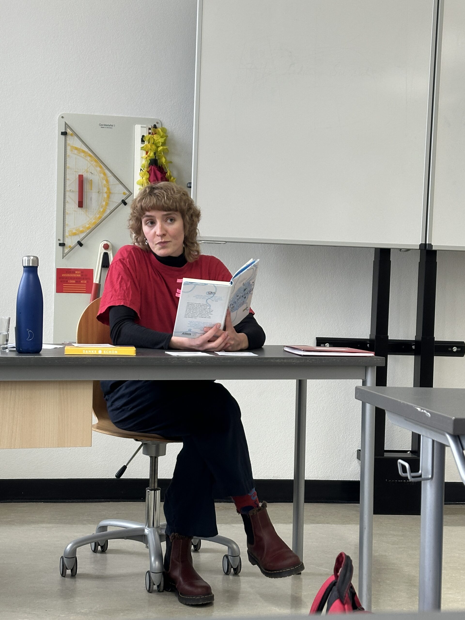 Eine Frau mit lockigem Haar, rotem Shirt, sitzt lächelnd an einem Tisch; hält ein Buch.