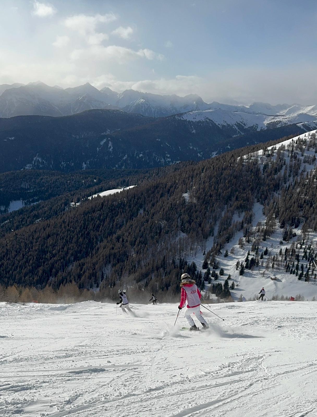 Eine Person in rosa Jacke fährt Ski auf verschneitem Berg mit bewaldetem Tal im Hintergrund.