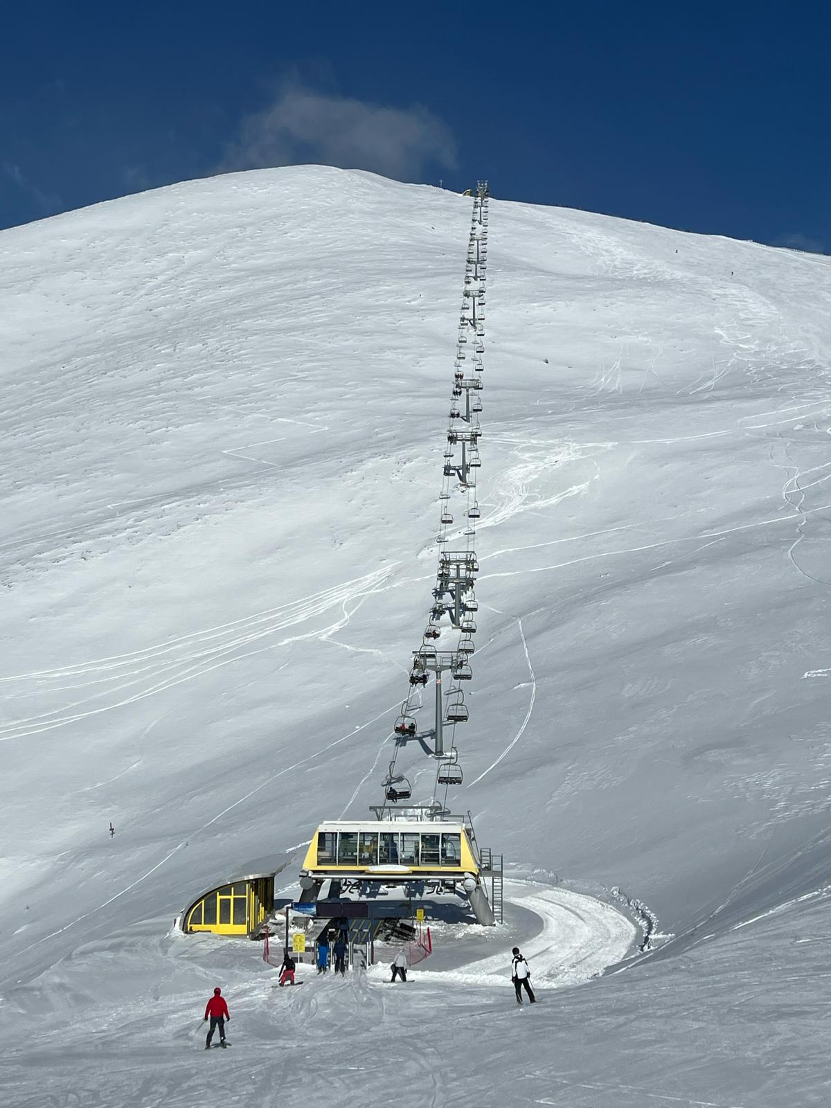 Skilift in Schneelandschaft, weiße Pisten mit blauen Himmel; Menschen in bunter Winterkleidung.